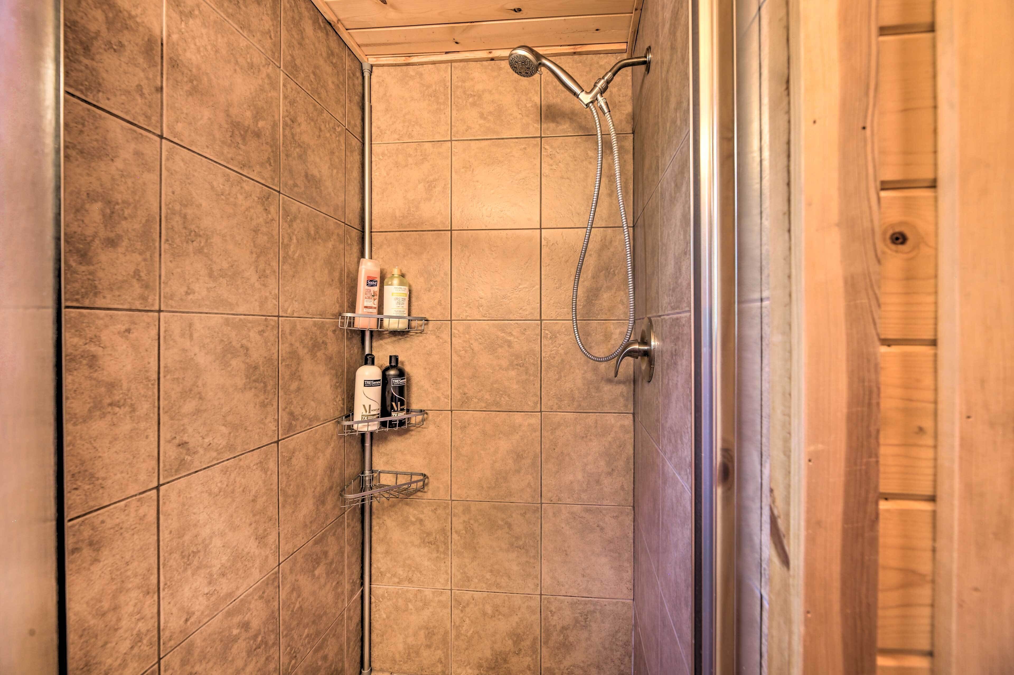 A shower with a handheld showerhead and a wall-mounted faucet, tiled walls, and a wooden door frame.