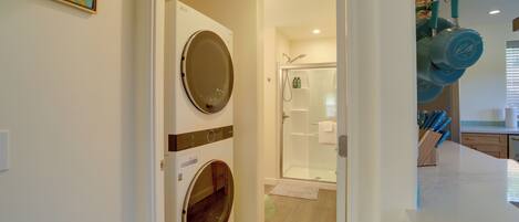 A laundry room with a front-loading washer and dryer, a glass shower enclosure, and a kitchen area with blue kettles and knives.