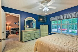 A bedroom with a bed, dresser, TV, and a window with a view of a red car and trees.