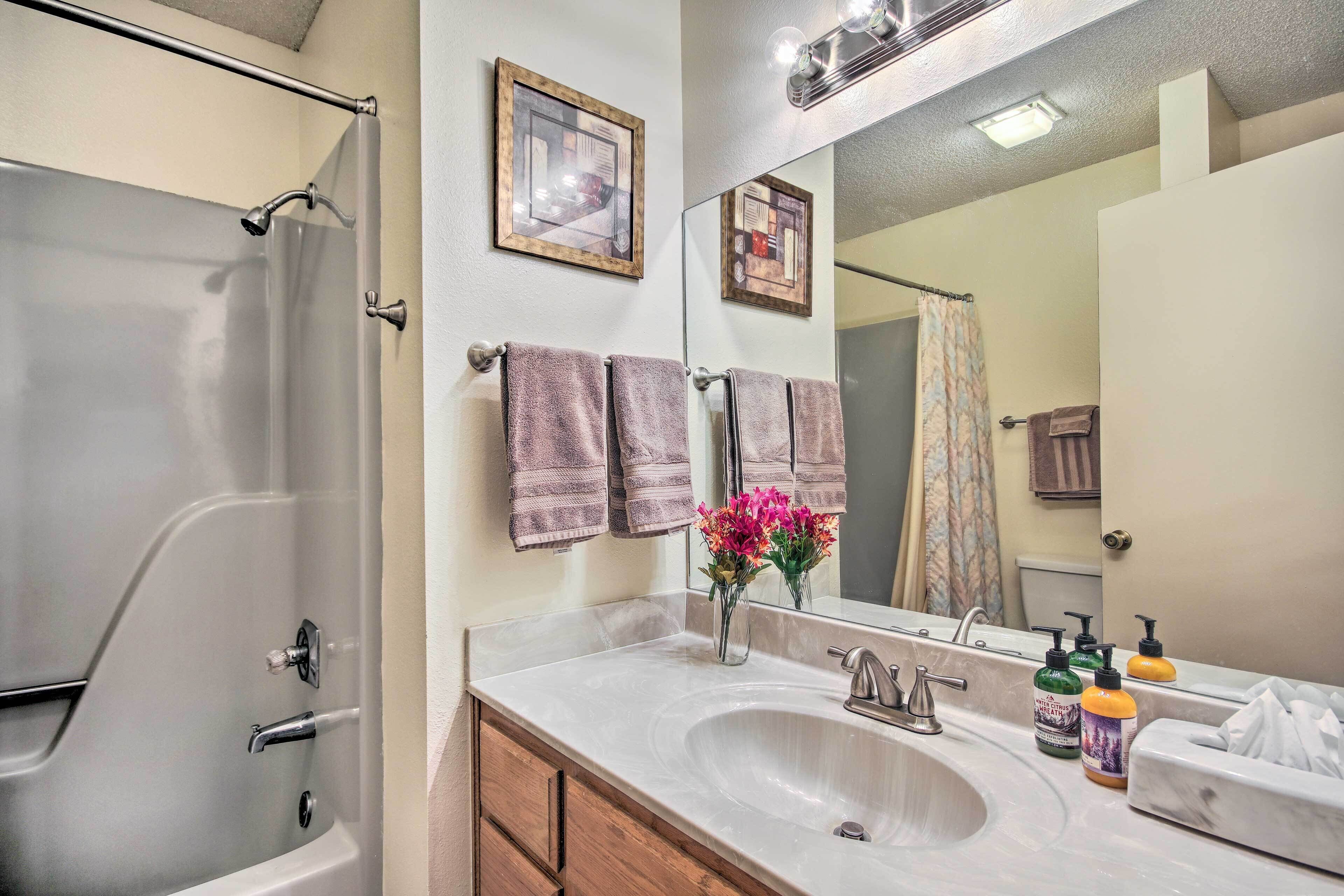 A bathroom with a bathtub, a sink with two faucets, and a mirror above the sink. There are towels hanging on the wall and a vase with flowers on the sink counter.