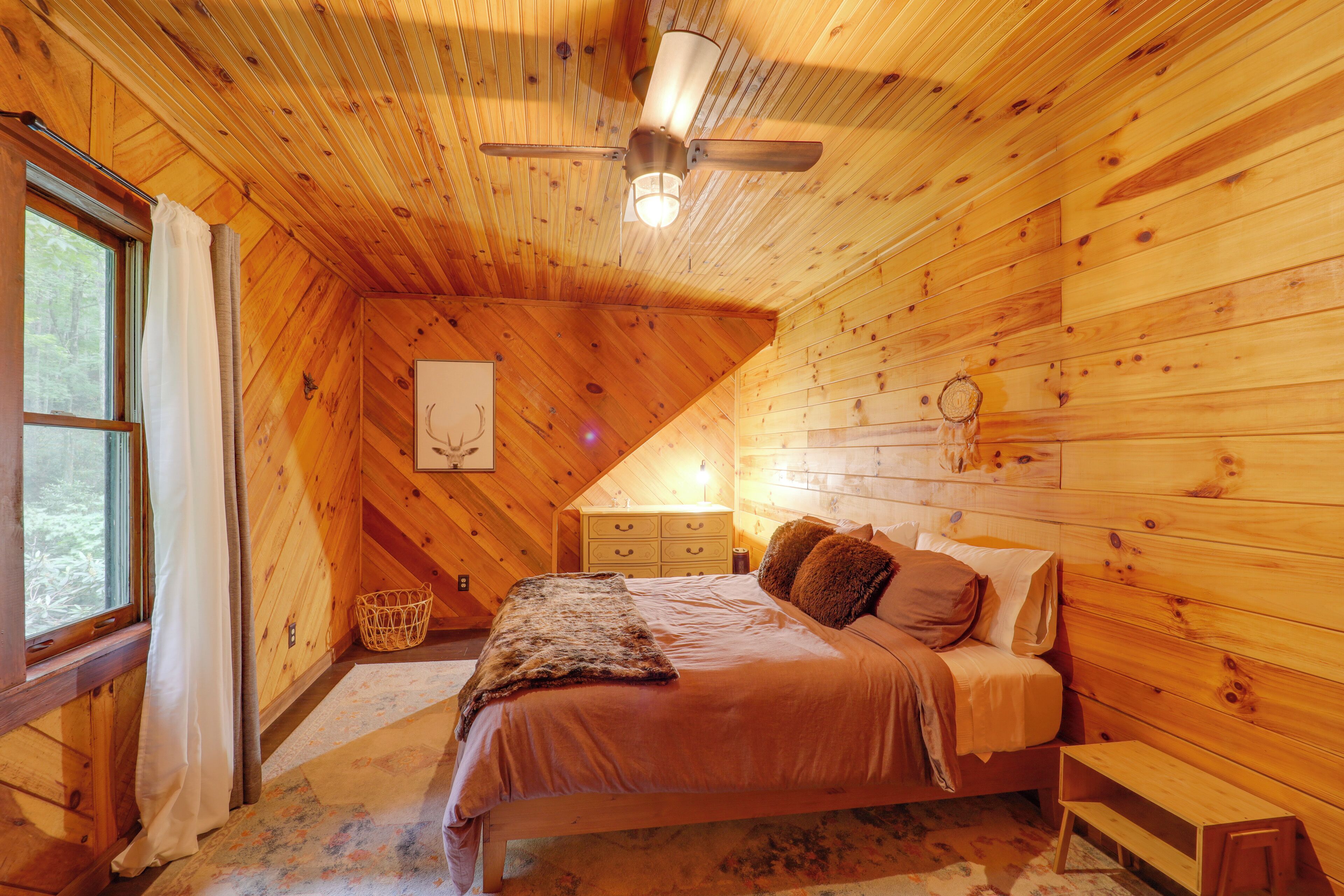 Een gezellige slaapkamer met een houten plafond, een bed met dekens en kussens, een nachtkastje, een dressoir en een raam met gordijnen.