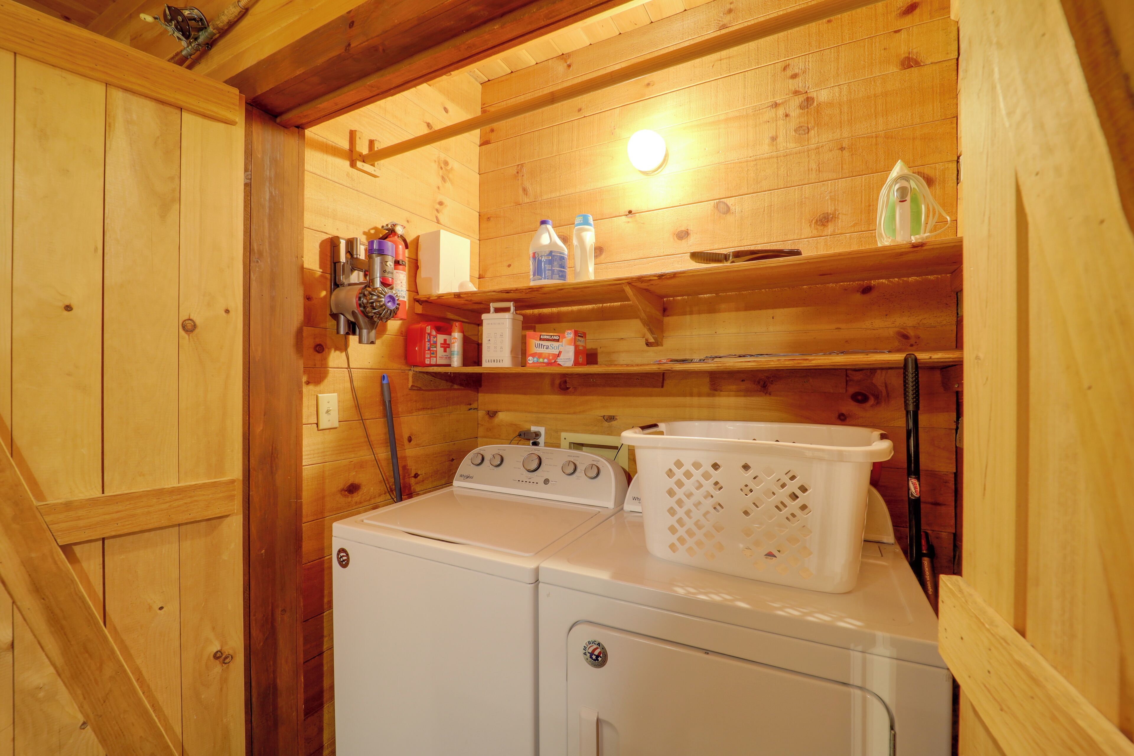 Un espace réduit dédié à la buanderie, comprenant un lave-linge et un sèche-linge, des étagères en bois où sont rangés divers objets, et un panier.