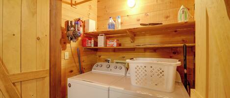 Un espace réduit dédié à la buanderie, comprenant un lave-linge et un sèche-linge, des étagères en bois où sont rangés divers objets, et un panier.