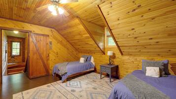A cozy attic bedroom with wooden walls and a sloped ceiling. There are two beds with matching bedding, a small bedside table with a lamp, and a patterned rug on the floor.