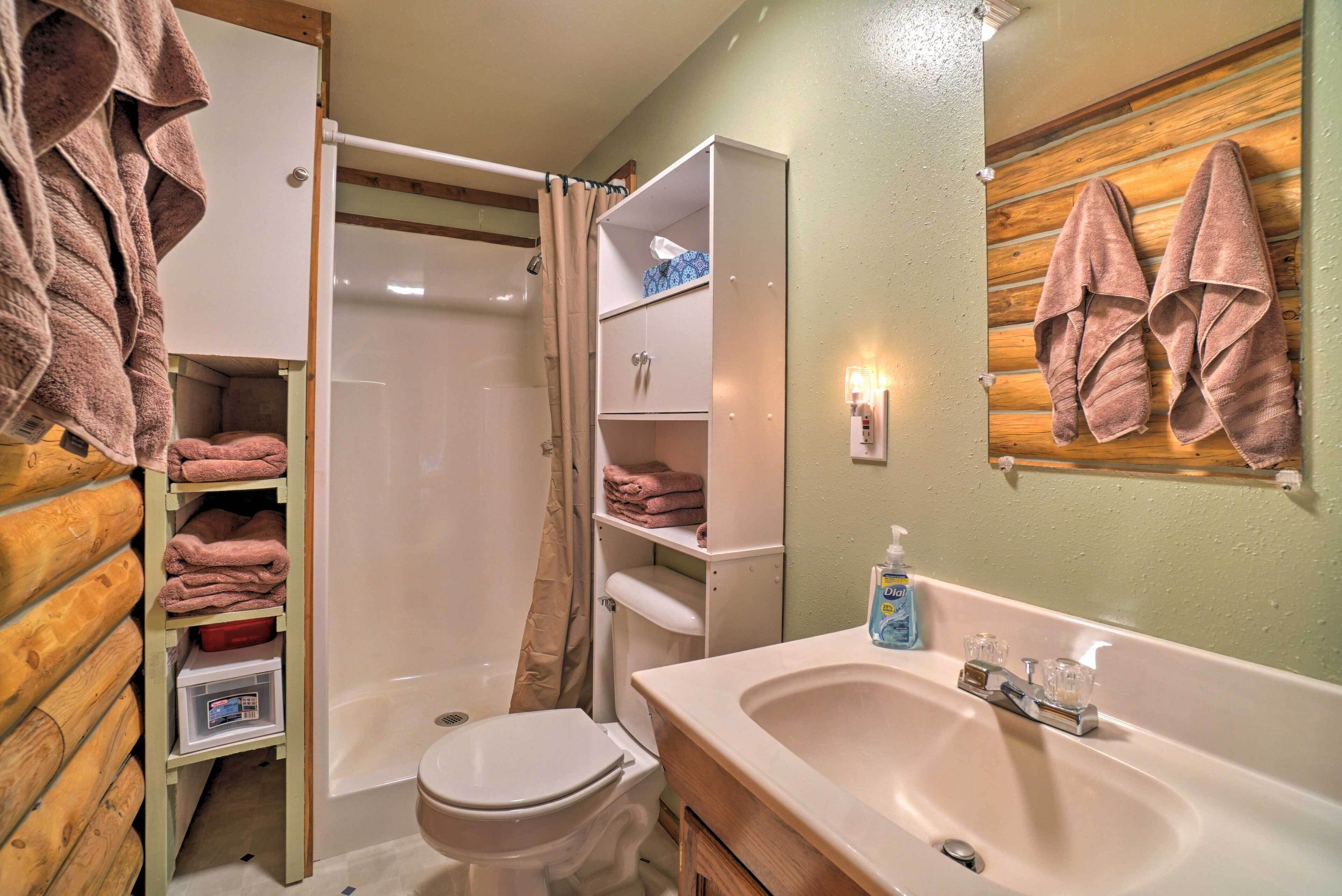 A bathroom with a shower, toilet, and sink. There are towels hanging on the wall and shelves with folded towels.