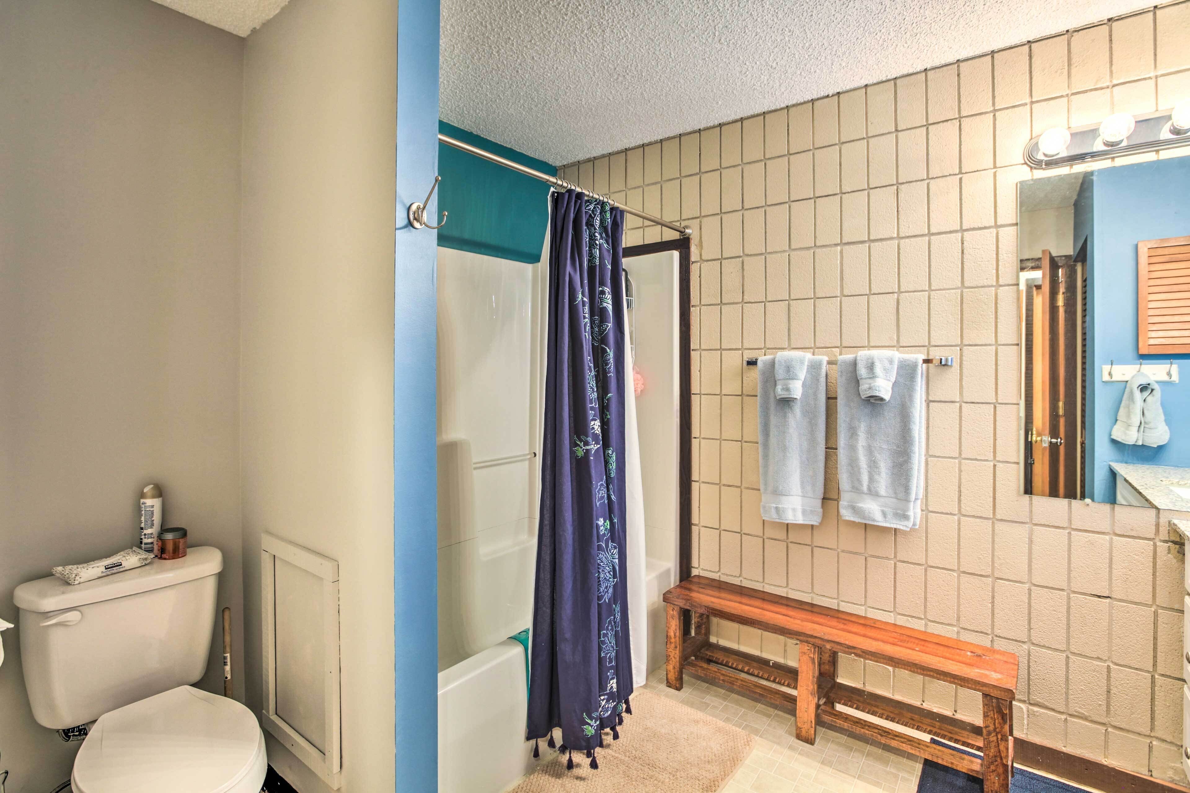 Een badkamer met toilet, douche met gordijn, een zitbankje, betegelde muur, spiegel en handdoekhouder.
