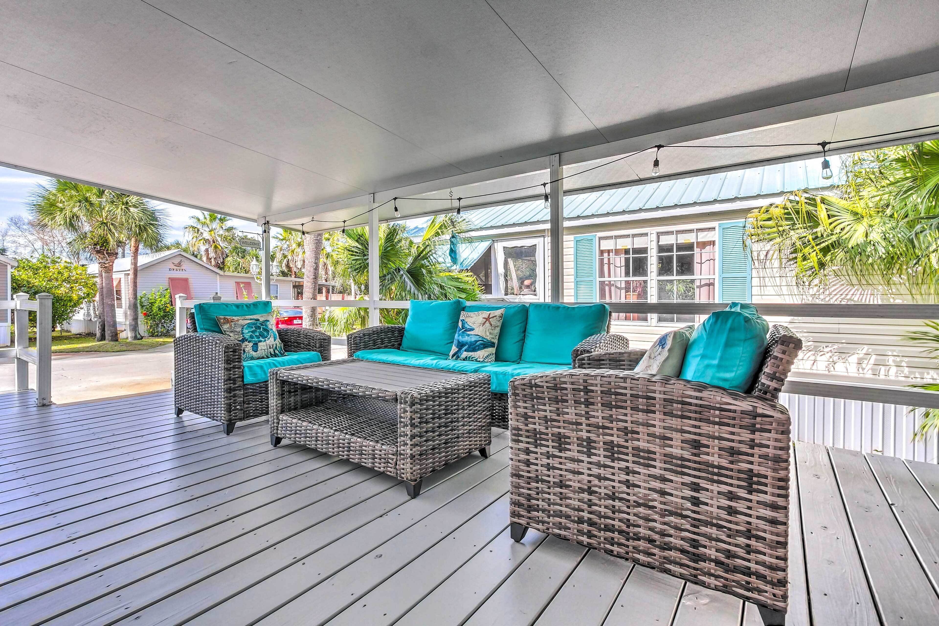 A covered outdoor seating area with wicker furniture and teal cushions, overlooking a residential area with palm trees and a house with a blue roof.