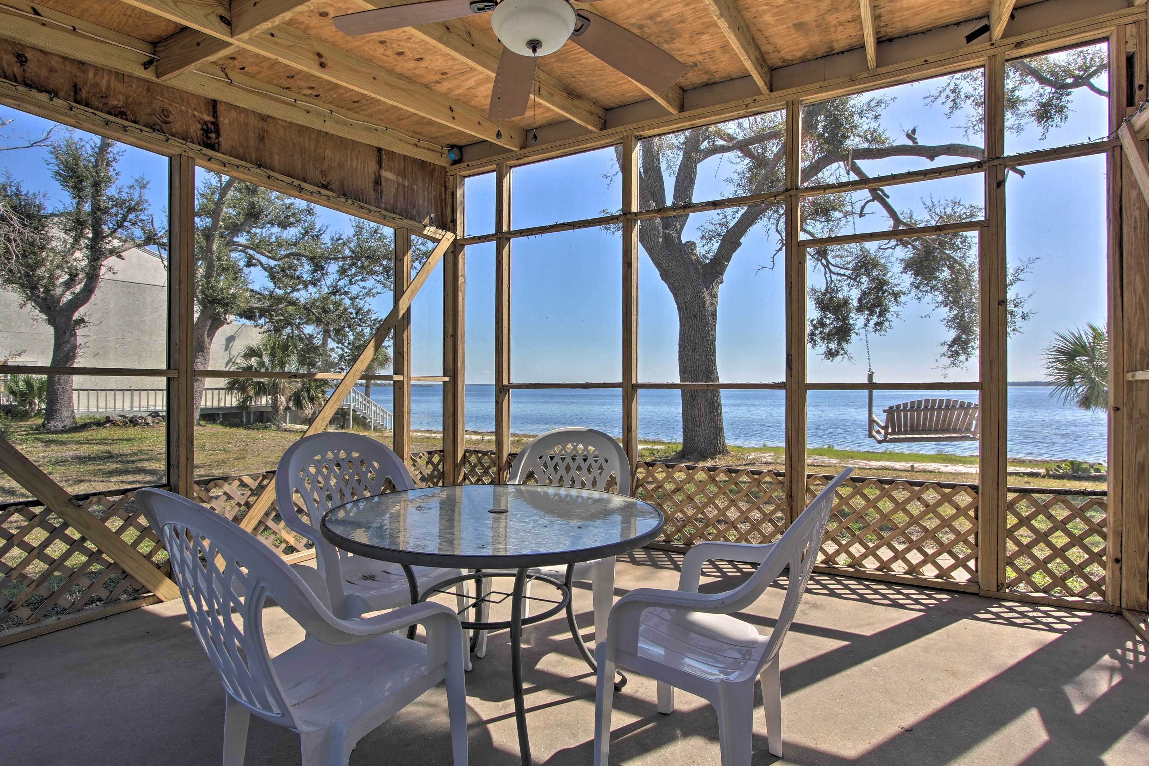 A covered patio with a round glass table and white plastic chairs, overlooking a beach and ocean.