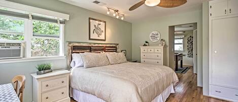 A bedroom with a large bed, a ceiling fan, a window with blinds, a bedside table with a lamp, and a wardrobe.
