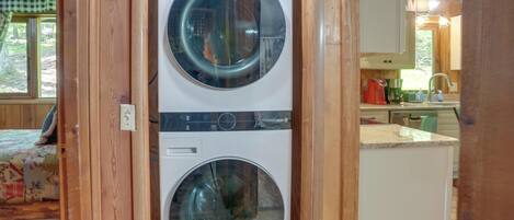 A laundry room with two front-loading washing machines, wooden walls, and a kitchen area visible through an open door.