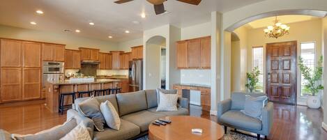 A spacious living room with a large sofa, a coffee table, and a fireplace. There is a kitchen area with wooden cabinets and a dining table.
