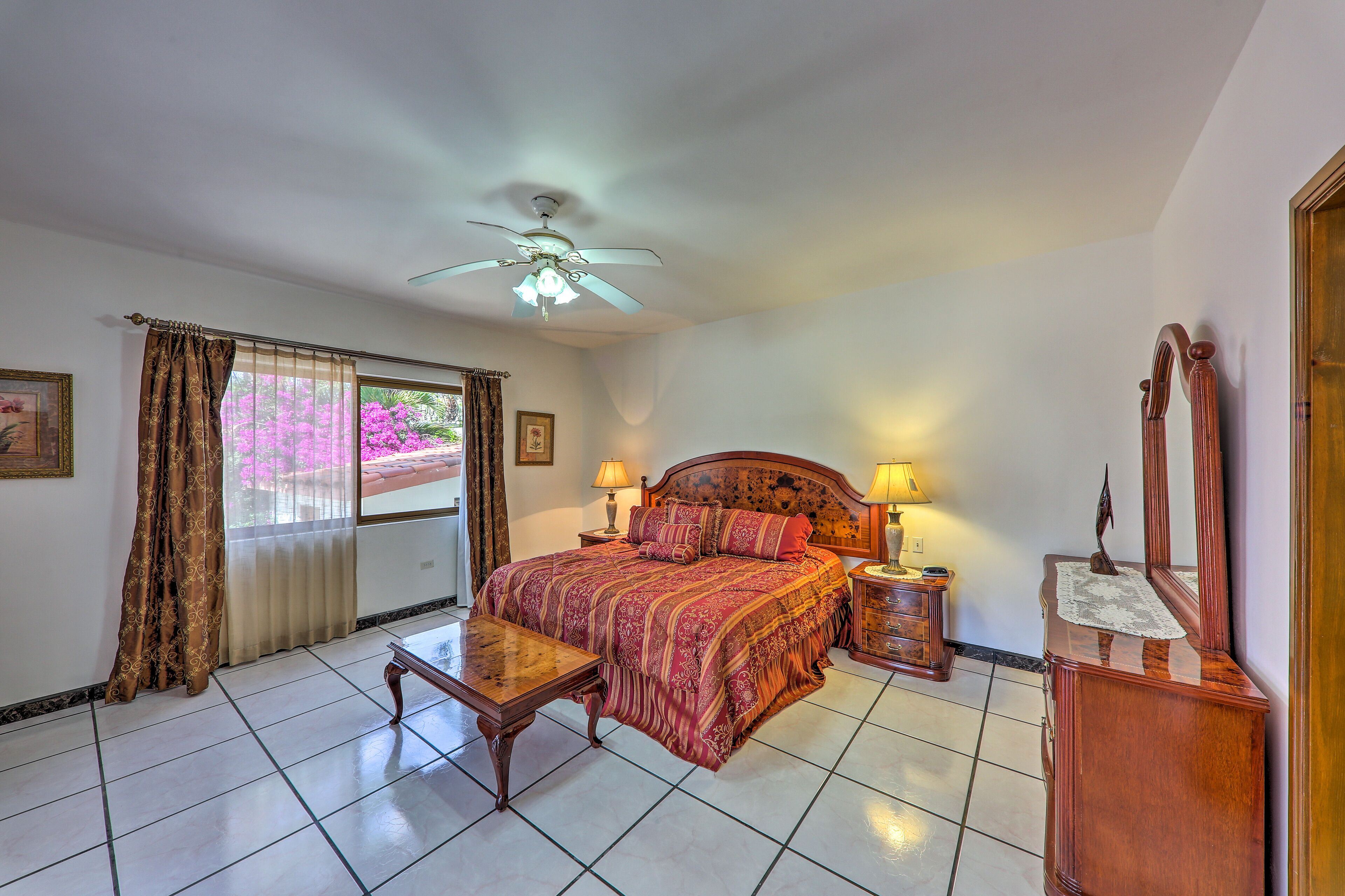 A bedroom with a bed, bedside tables, a ceiling fan, a window with curtains, and a wooden dresser.