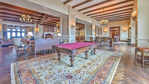 A spacious room with a pool table, wooden beams, chandeliers, and a large patterned rug.