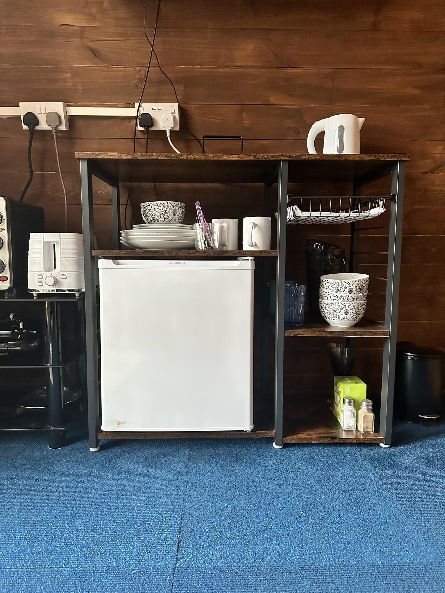 Et kompakt tekøkken med et hvidt køleskab, en lille toaster og en kaffemaskine. Der er hylder med service og en elkedel.