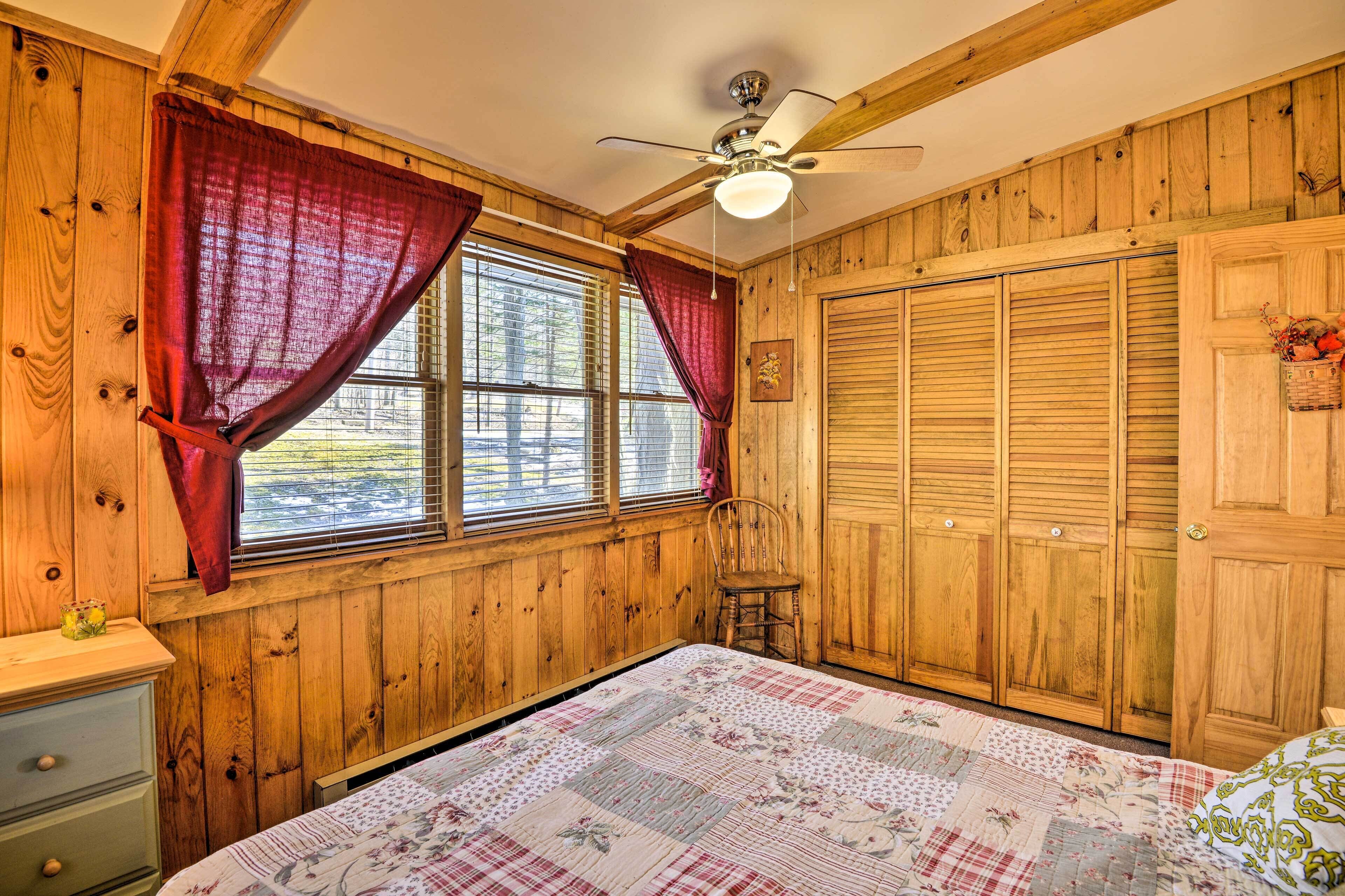 A wooden-paneled room with a bed, a chair, a ceiling fan, and a window with curtains.