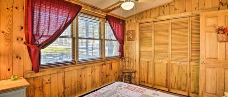 A wooden-paneled room with a bed, a chair, a ceiling fan, and a window with curtains.