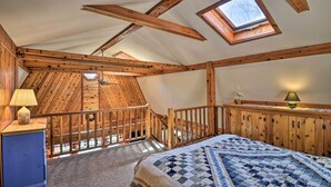 Une chambre avec un plafond en pente, des poutres en bois, un puits de lumière, un lit recouvert d’une courtepointe, une table de chevet avec une lampe et une commode.