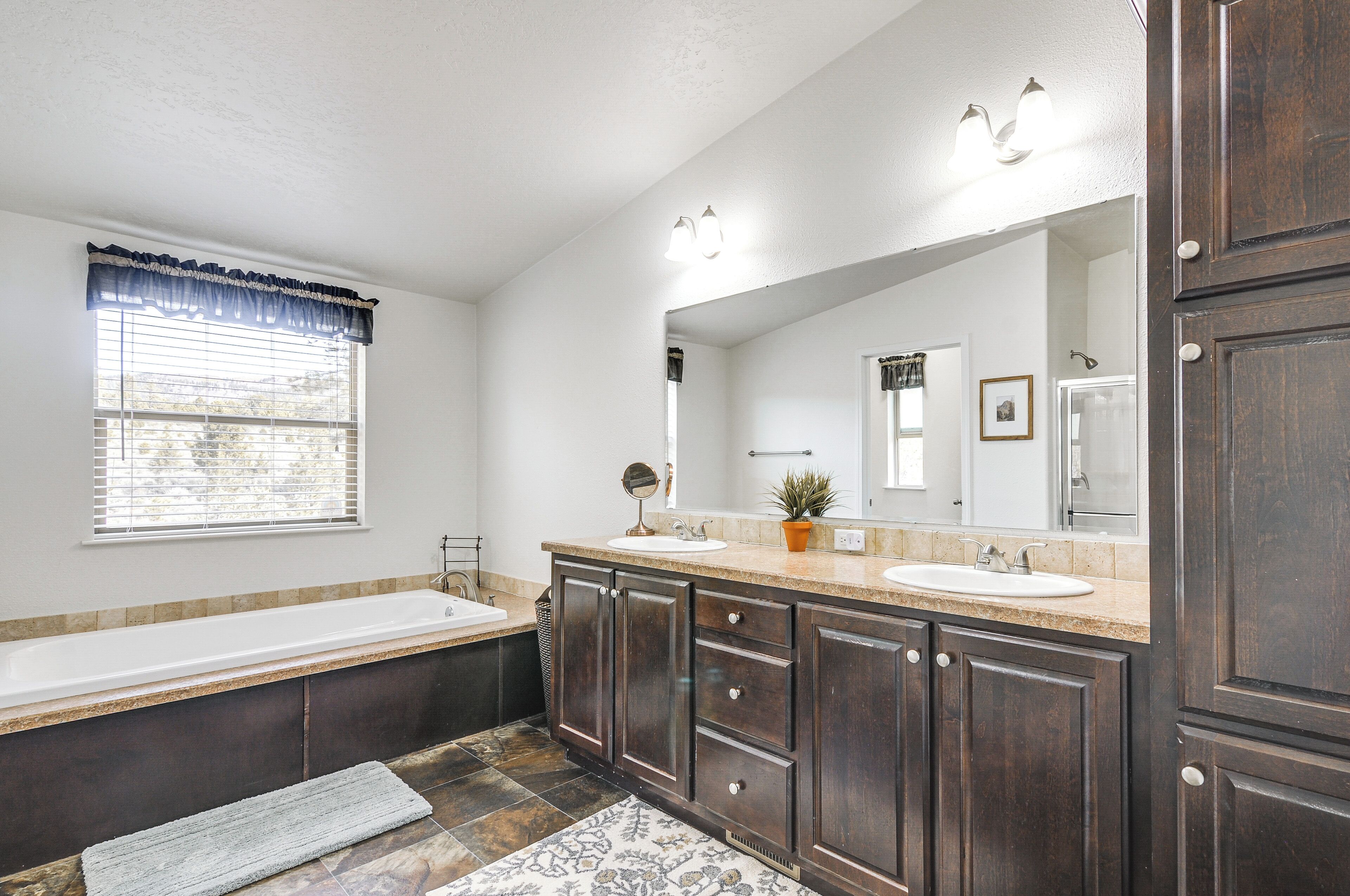 A bathroom with a bathtub, two sinks, dark wooden cabinets, a large mirror, and a window with blinds.
