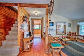 Interior de cabana de madeira com escada, área de refeições com mesa e cadeiras, e televisão fixada na parede.