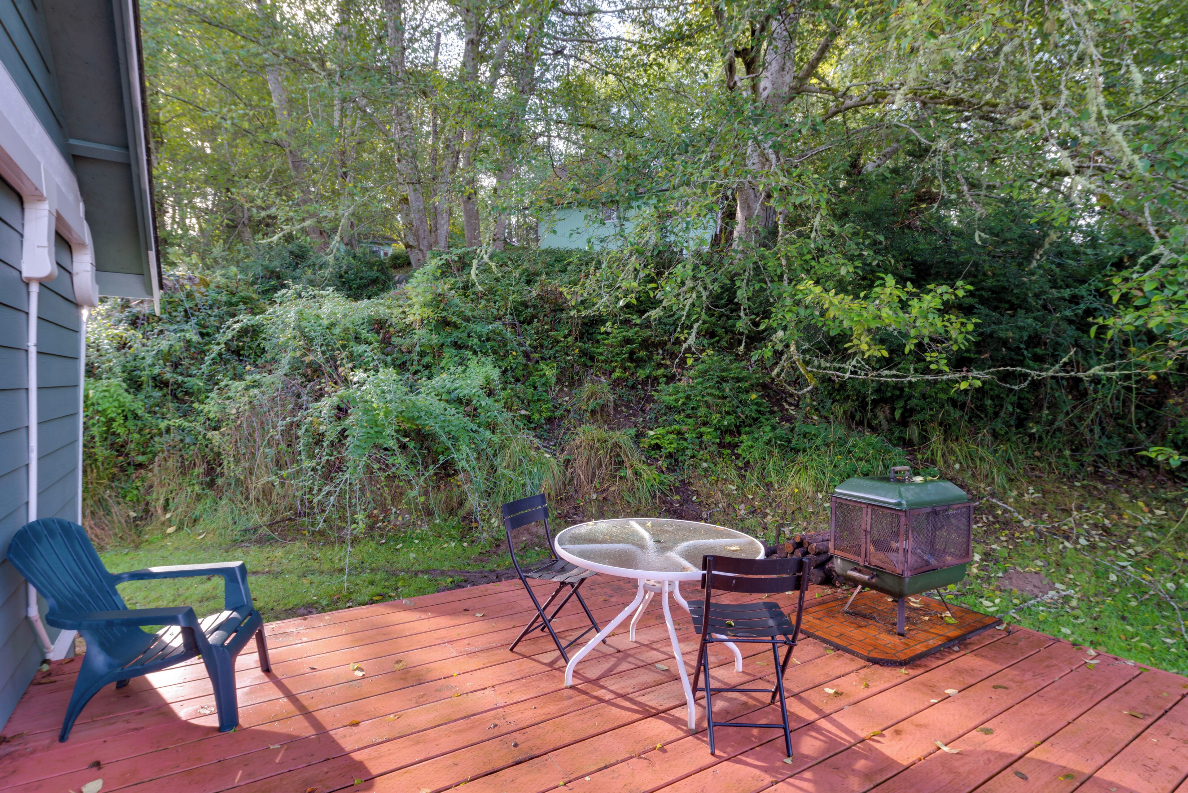 A wooden deck with a table and chairs, surrounded by dense foliage and a green building in the background.