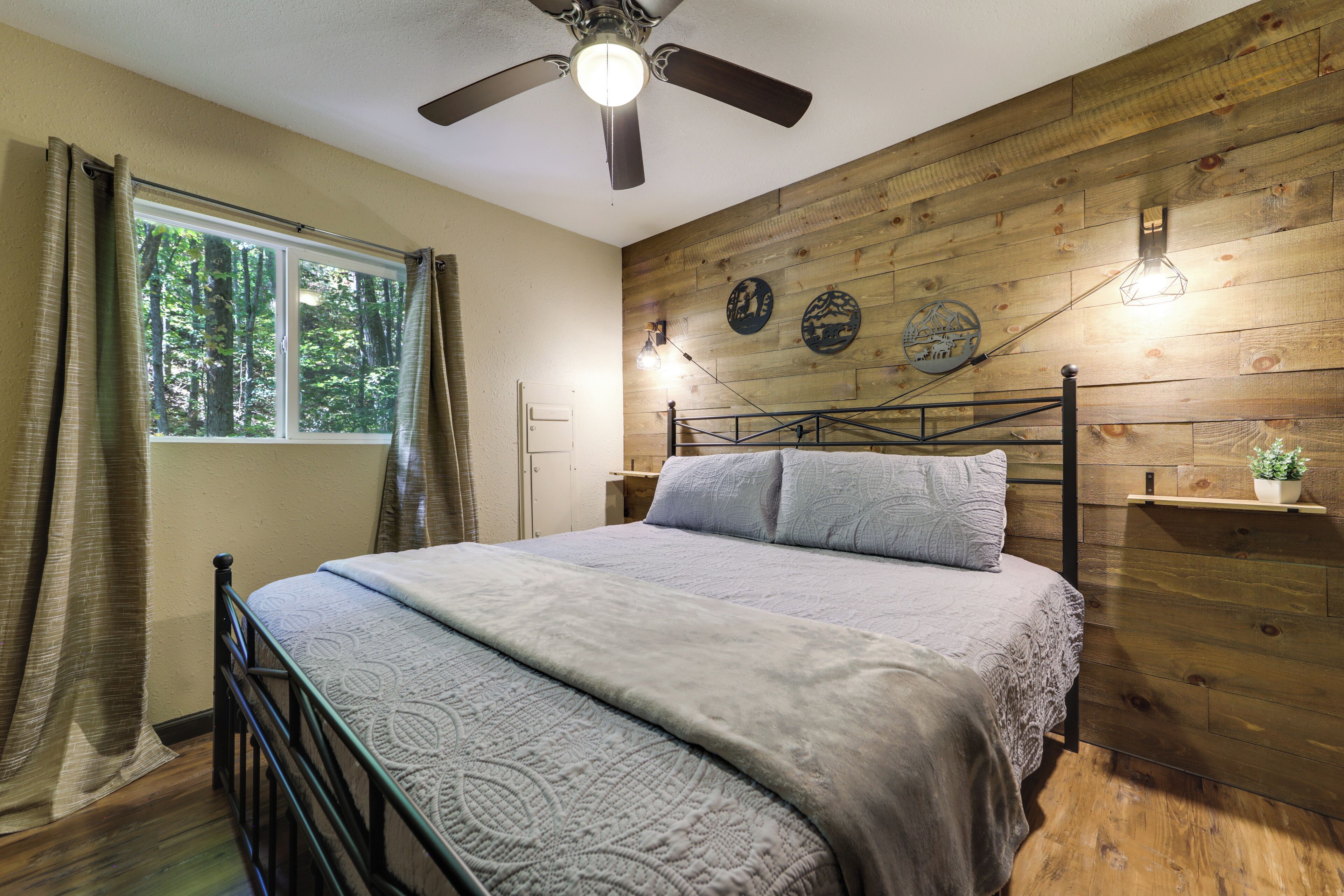Une chambre avec un mur en bois, un lit avec une tête de lit, un ventilateur de plafond, une fenêtre avec des rideaux et des lampes fixées au mur.