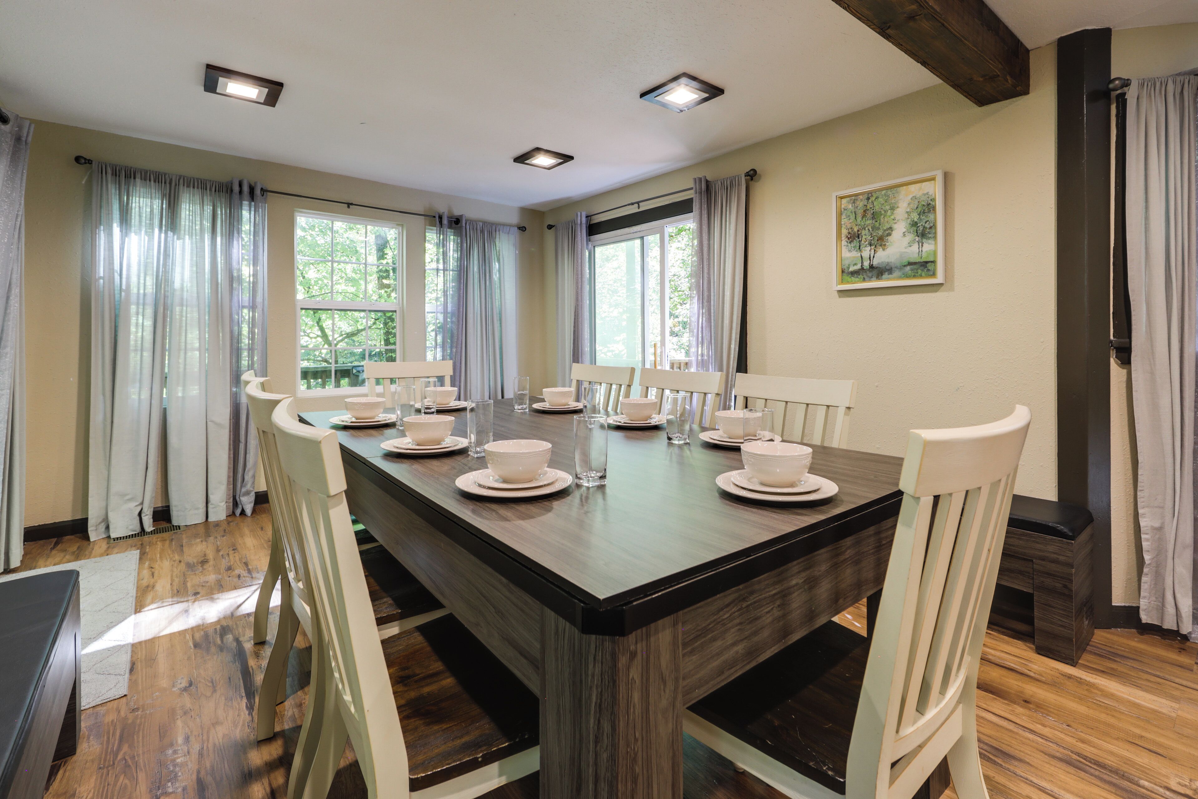 A dining area with a long table set for four, surrounded by white chairs, with a painting on the wall and a window with curtains.