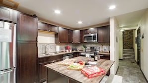 A modern kitchen with dark wood cabinets, stainless steel appliances, and a central island with fruit and a pizza box.