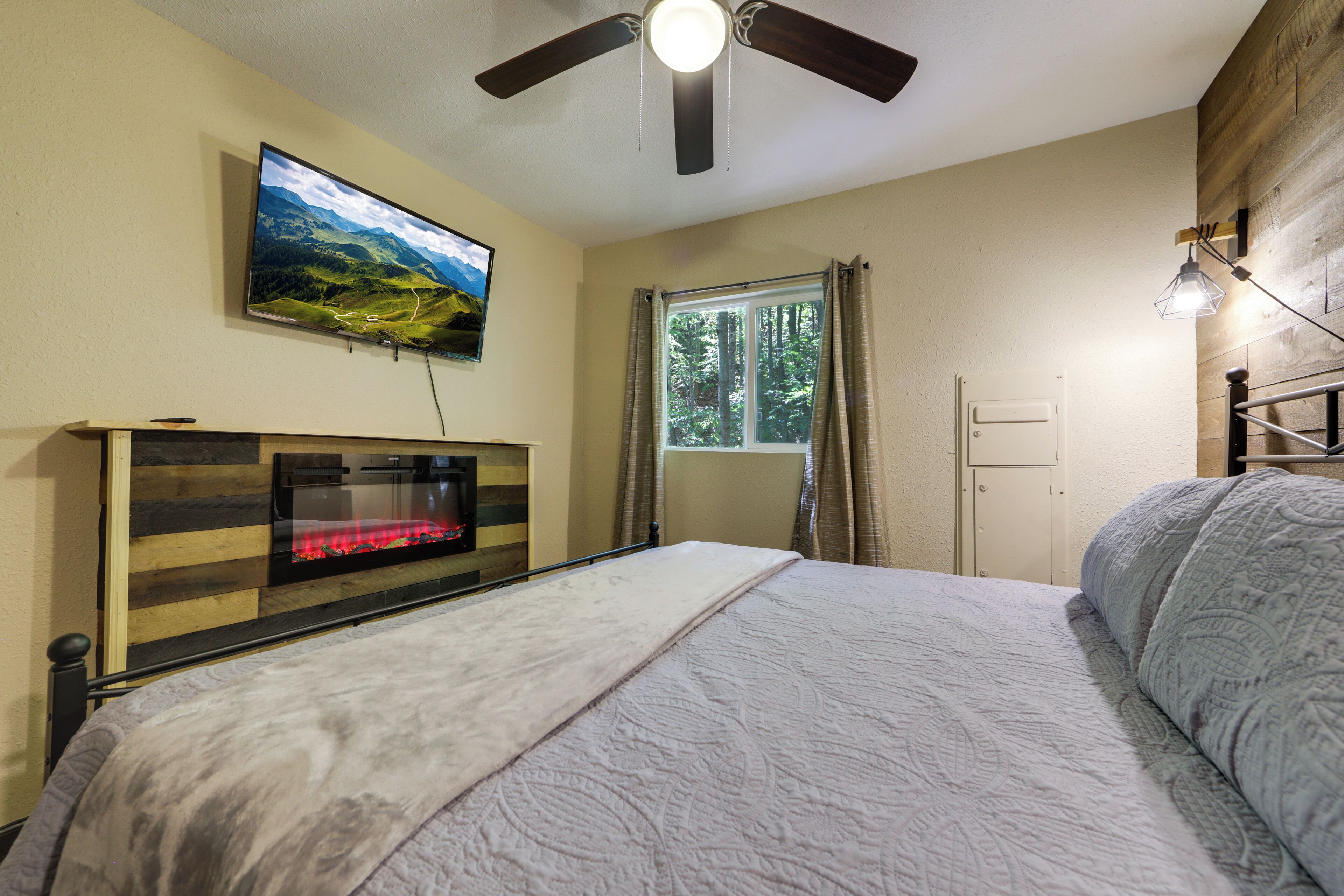 A bedroom with a bed, a television mounted on the wall, a window with curtains, and a ceiling fan.