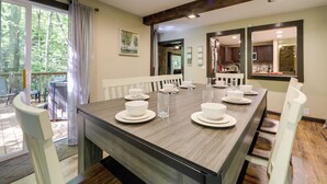 A dining area with a long table set for six, white chairs, and a view of the outdoors through a glass door.