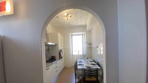 A narrow hallway leading to a dining area with a table set for four, a kitchen with white cabinets, and a window providing natural light. - Home Roman Center Neptune (Nettuno)