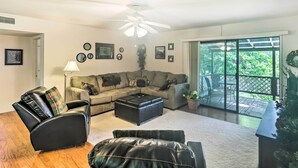 A living room with a sofa, a leather chair, a coffee table, and a ceiling fan. There is a large window with a view of greenery outside.