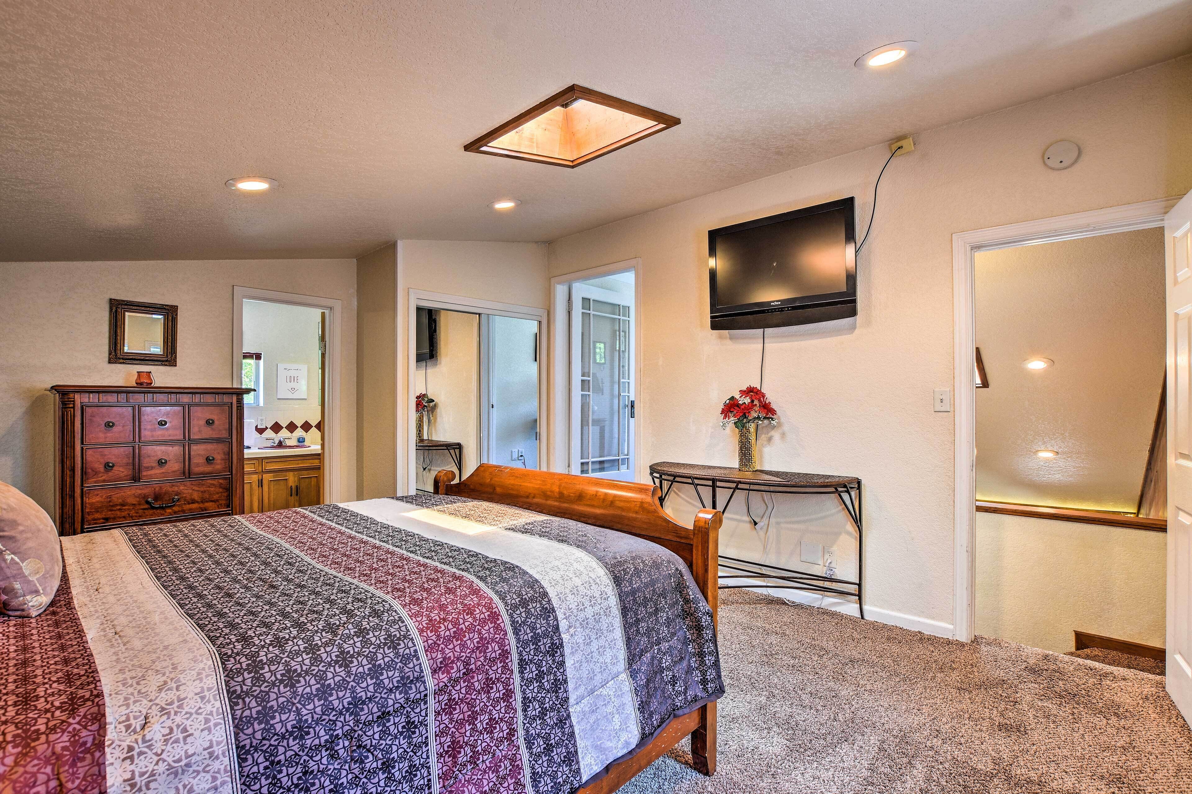 A bedroom with a large bed, a dresser, a TV mounted on the wall, and a door leading to another room.