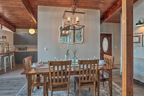 A dining area with a wooden table, chairs, and a chandelier. There is a kitchen in the background with cabinets and a microwave.