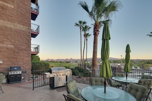 An outdoor patio with a grill, chairs, and umbrellas, overlooking a golf course and mountains.