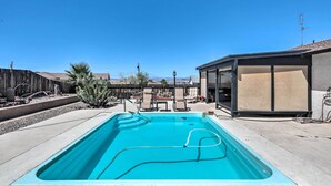 A rectangular pool with a clear blue waterline, surrounded by a concrete deck. There are lounge chairs and a small table near the pool. A wooden fence and desert plants are visible in the background.