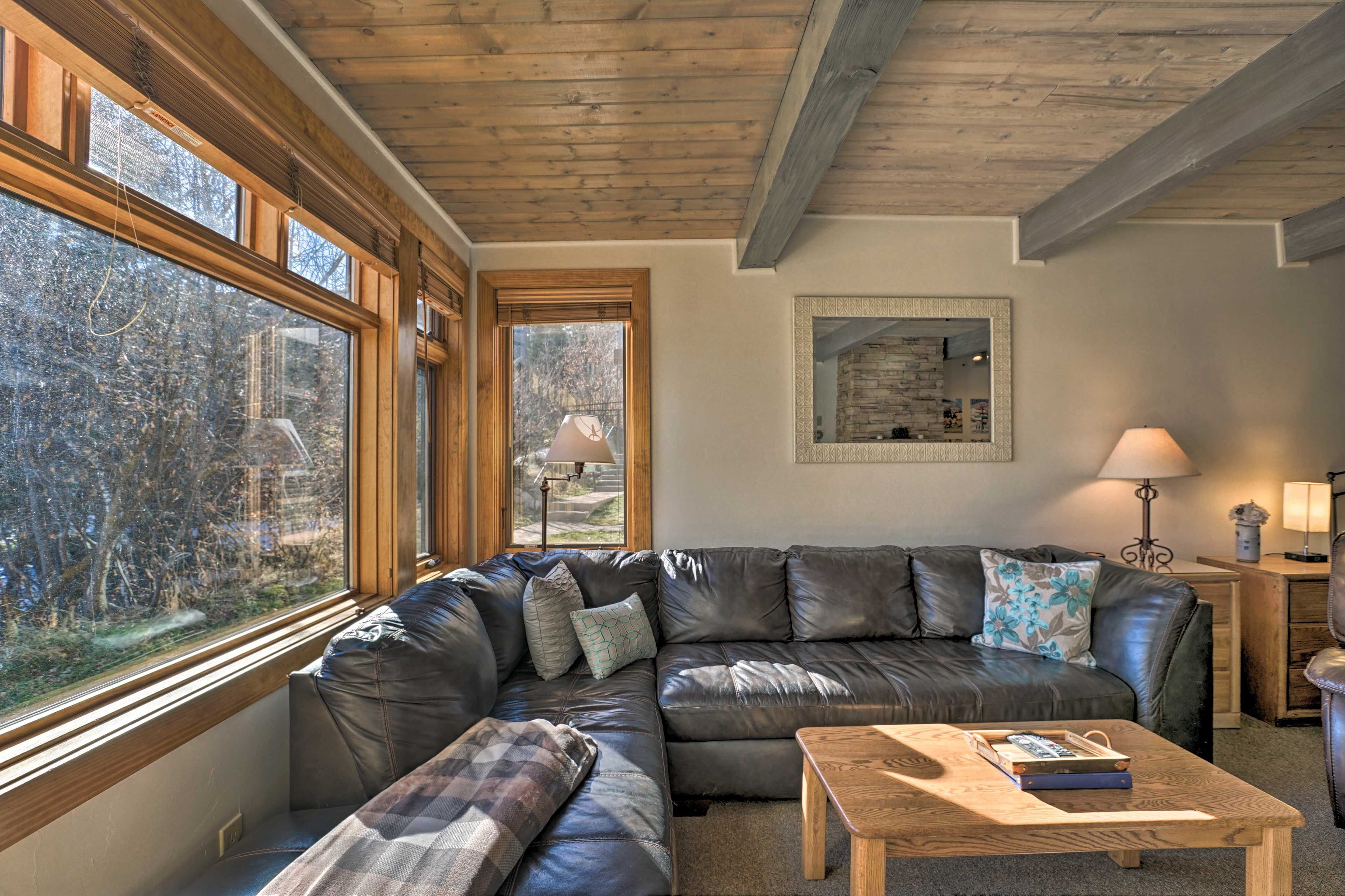 Ein gemütliches Wohnzimmer mit einer Eckcouch, einem Holztisch und einem großen Fenster mit Blick auf Bäume.