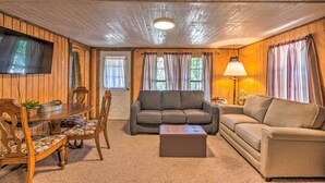 Ein gemütliches Wohnzimmer mit Holzbalkendecke, einem Flachbildfernseher, einem grauen Sofa, einem beigefarbenen Sofa, einem Holzesstisch mit Stühlen und einem kleinen Tisch mit einer Lampe.