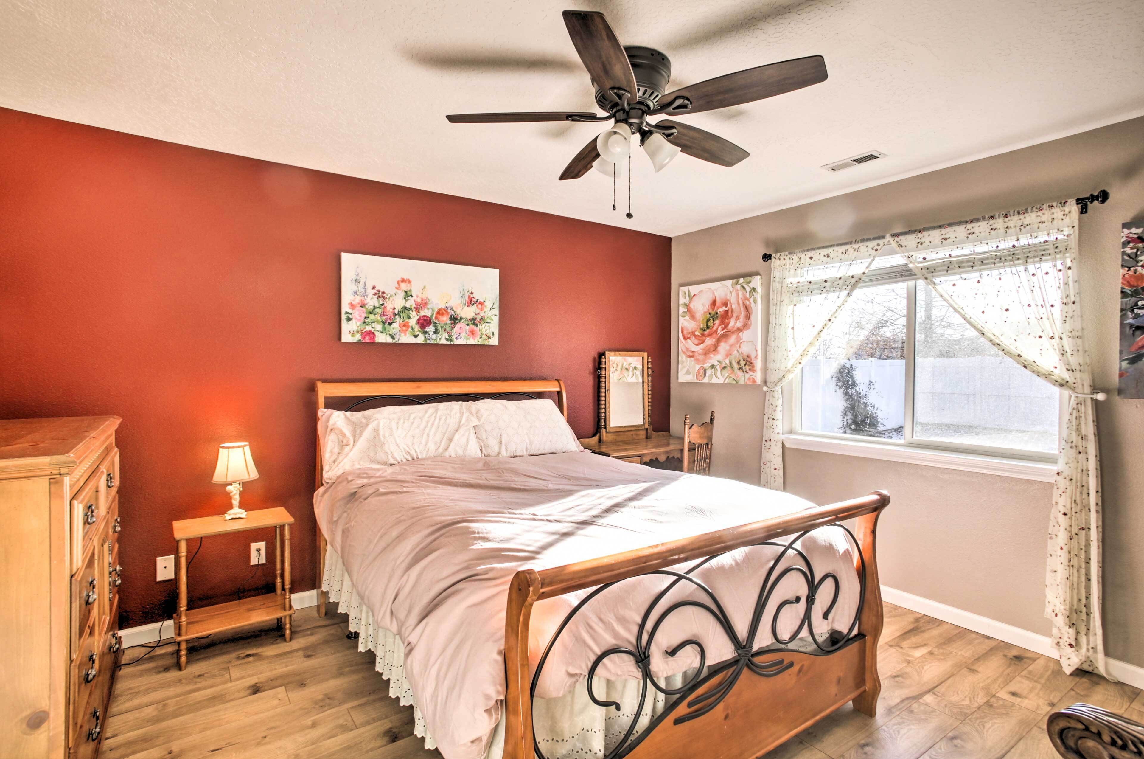 A bedroom with a wooden bed, a ceiling fan, a nightstand with a lamp, and floral artwork on the walls.