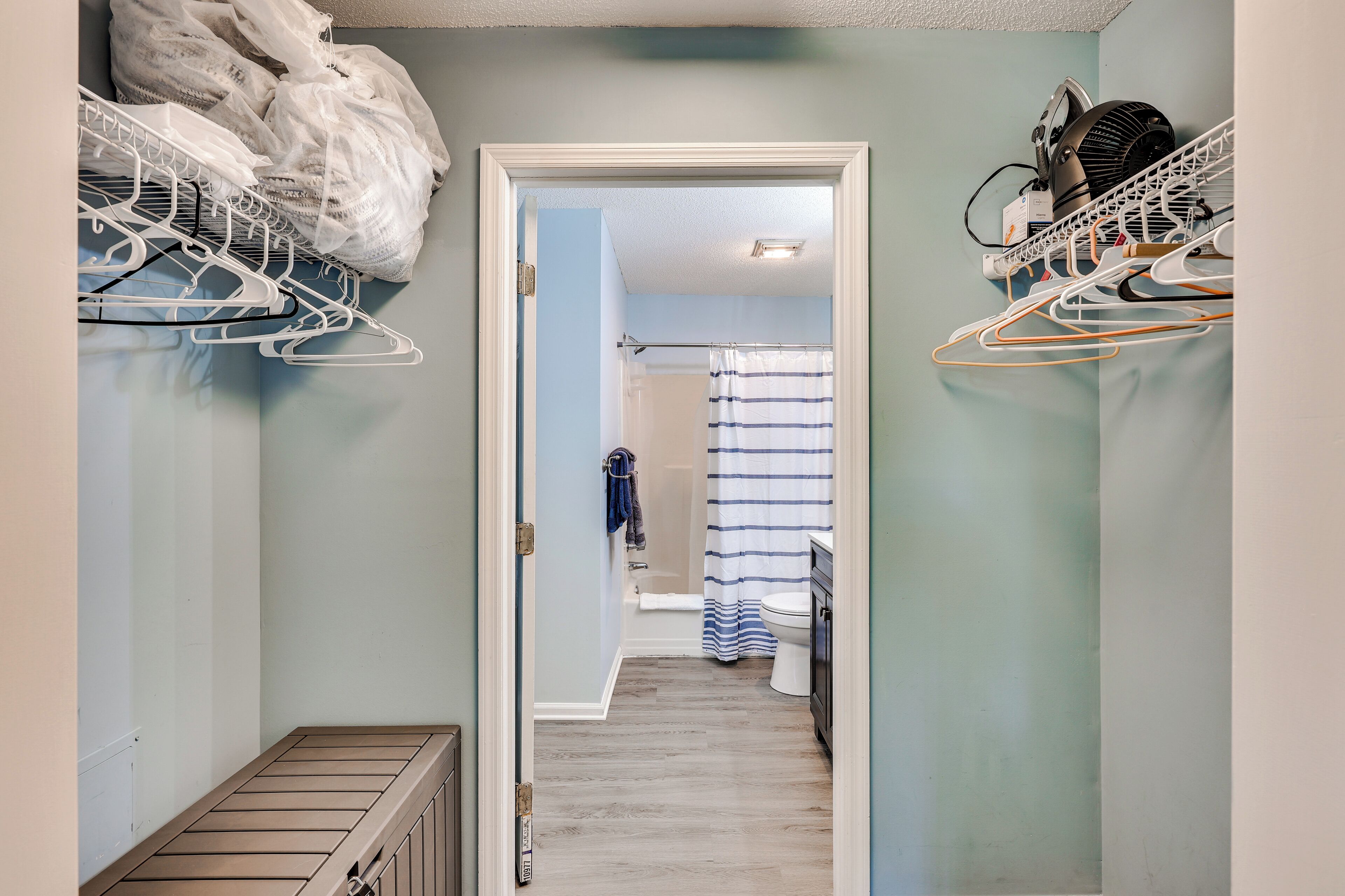 Un couloir étroit avec un radiateur, une porte blanche donnant sur une salle de bain avec un rideau de douche, et un coin buanderie avec des vêtements suspendus et un ventilateur.