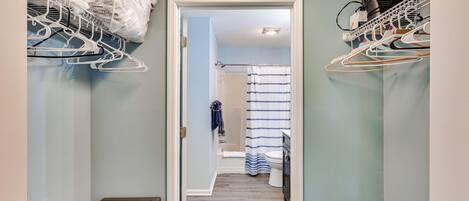 Un couloir étroit avec un radiateur, une porte blanche donnant sur une salle de bain avec un rideau de douche, et un coin buanderie avec des vêtements suspendus et un ventilateur.