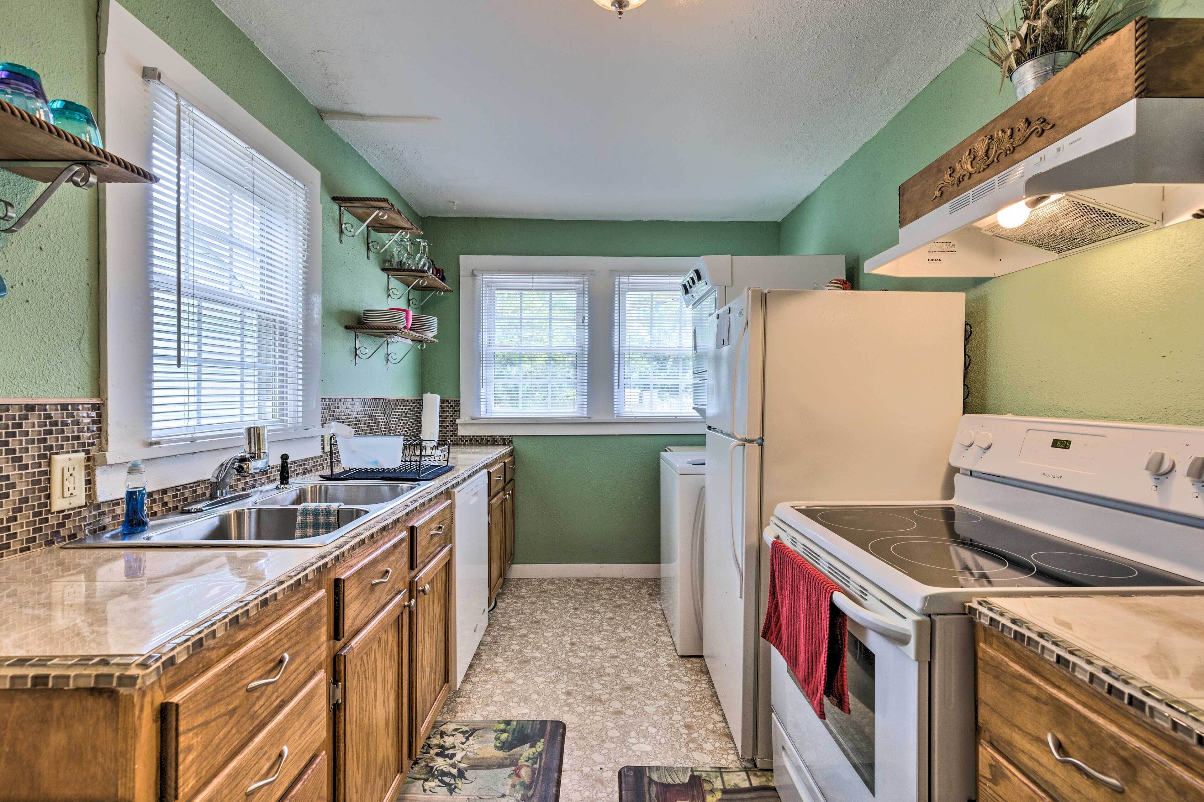 A compact kitchen with a refrigerator, stove, and sink. There are two windows with blinds, and the walls are painted green.