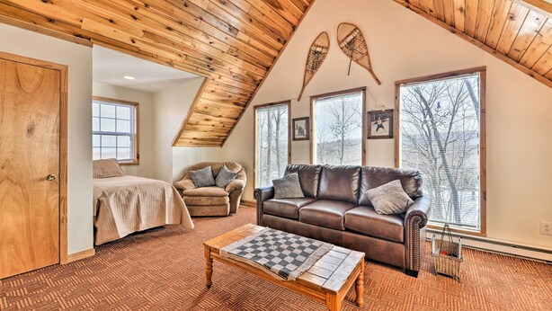 A cozy loft bedroom with a sloped ceiling, a large window, a bed, a brown leather sofa, and a wooden coffee table. - Ski-in/ski-out Ghent Cabin w/ Mountain Views! (Ghent)