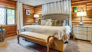 A log cabin bedroom with a large bed, a bench, a nightstand, and a window with curtains. - Lake Lure Home w/ Deck: At Lake Lure & Golf! (Lake Lure)
