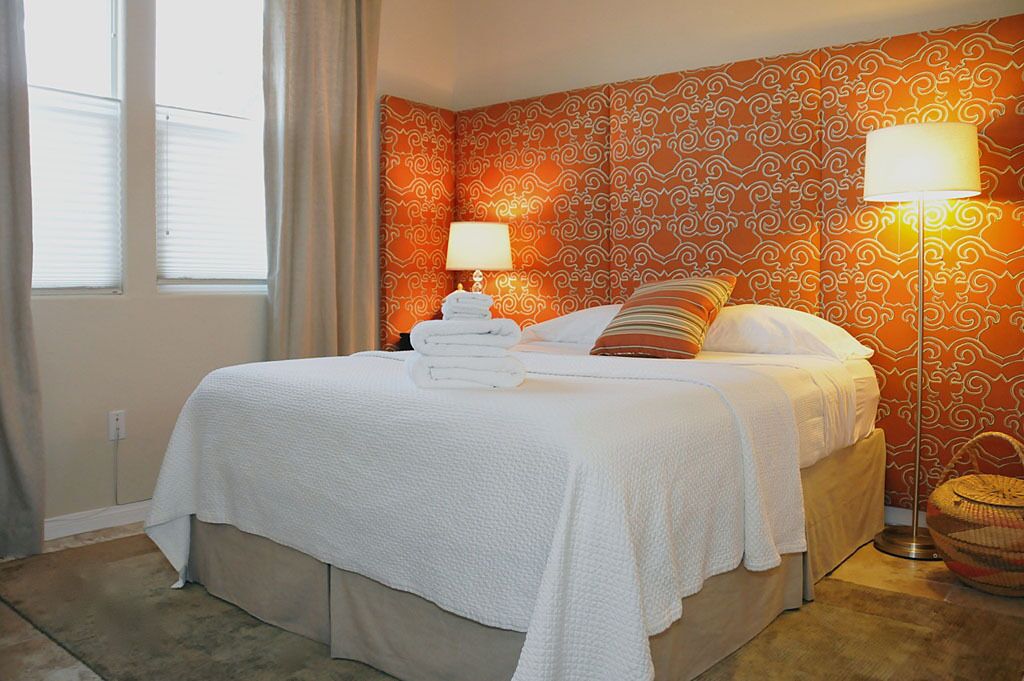 A neatly made bed with white linens, two bedside lamps, a patterned orange wall, and a window with curtains.