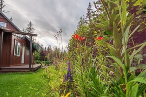 A wooden cabin with a porch, surrounded by lush greenery and various flowering plants. - Secluded Seward Home: Patio, 2 Mi to Kenai Fjords! (Seward)
