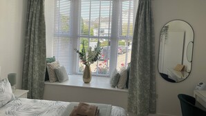 A bedroom with a bed, a window with curtains, a vase with flowers, a mirror, and a bedside table. - Swn Y Mor / Sound of the Sea (Criccieth)