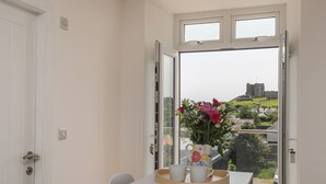 A room with a table set for two, a vase with flowers, and a view of a castle through a large window. - Swn Y Mor / Sound of the Sea (Criccieth)