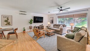 Un salon spacieux doté d’un plancher en bois, d’un ventilateur de plafond, d’une grande fenêtre avec des rideaux, d’un téléviseur à écran plat et d’un coin repas avec une table et des chaises.
