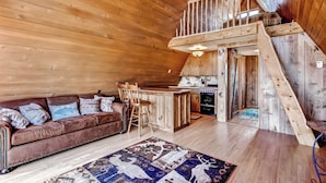 Un accogliente interno di una baita con soffitto in legno, un divano in pelle marrone, un tavolo da pranzo in legno con sedie e un angolo cottura.