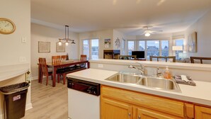 A modern kitchen with a dishwasher, sink, and wooden cabinets. There is a dining area with chairs and a table, and a view of the outside through large windows.
